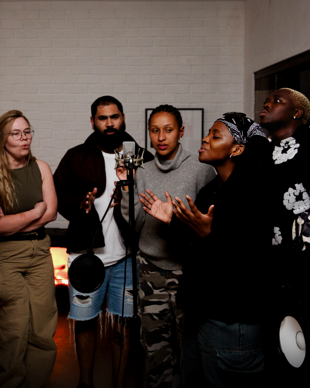 Fem personer står sammen indendørs med en mikrofon foran sig og ser ud til at synge eller tale mod en hvid murstensbaggrund.