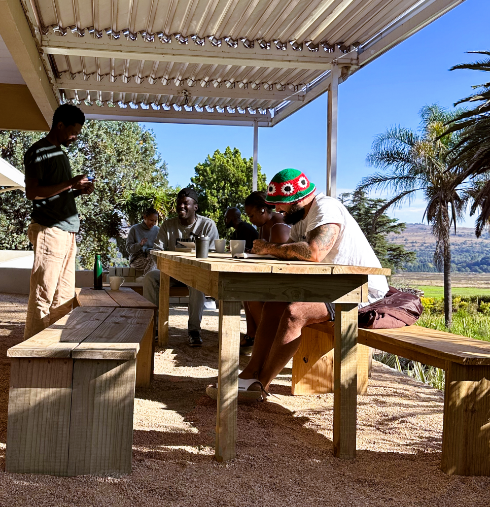 Seks personer sidder og står omkring et træbord under en pergola i det fri med træer, palmer og bakker i baggrunden på en solrig dag.