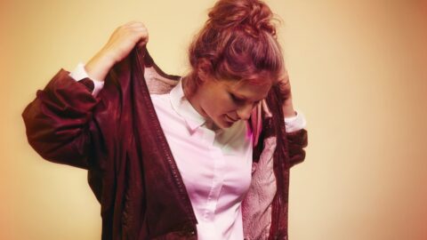 En kvinde med håret i en knold tager en brun jakke på over en hvid skjorte med knapper og kigger nedad mod en varm baggrund med gradienter.