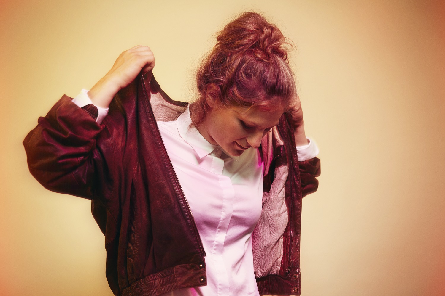 En kvinde med håret i en knold tager en brun jakke på over en hvid skjorte med knapper og kigger nedad mod en varm baggrund med gradienter.