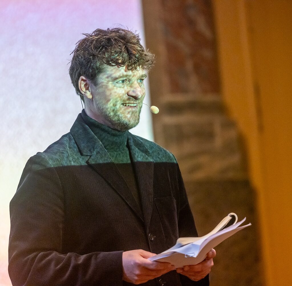 En mand iført sort jakke og headset-mikrofon holder papirer, mens han står foran en projiceret skærm.