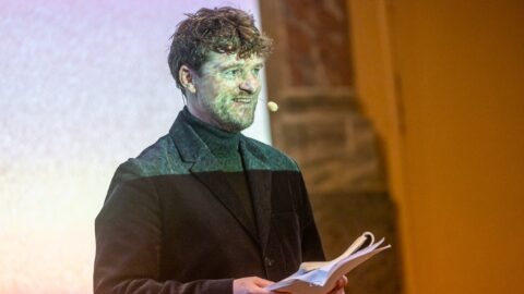 En mand iført sort jakke og headset-mikrofon holder papirer, mens han står foran en projiceret skærm.