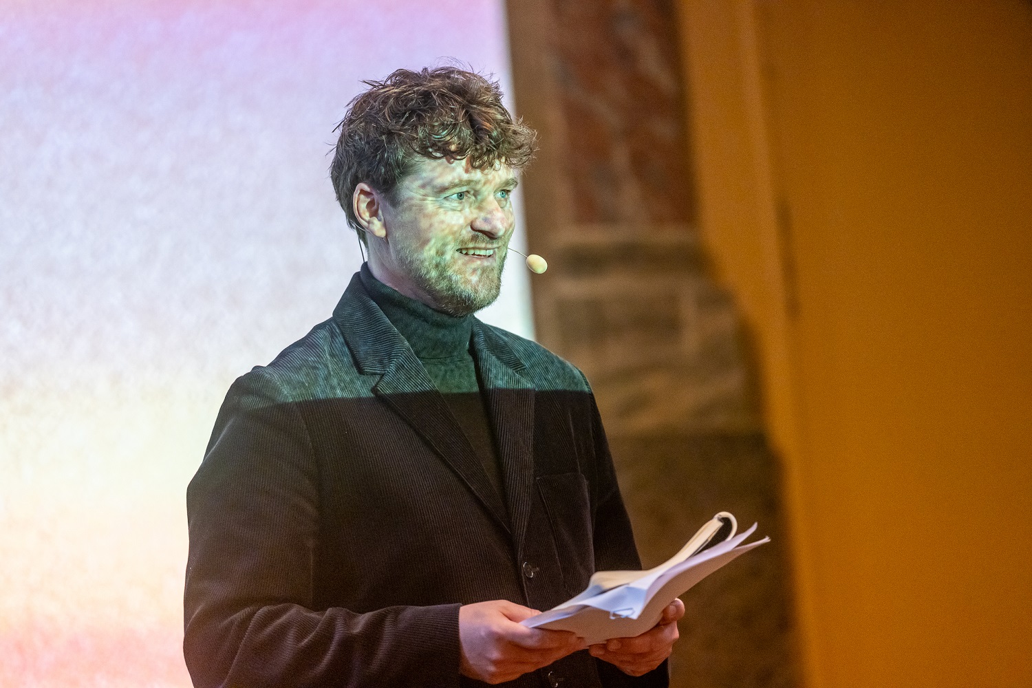 En mand iført sort jakke og headset-mikrofon holder papirer, mens han står foran en projiceret skærm.