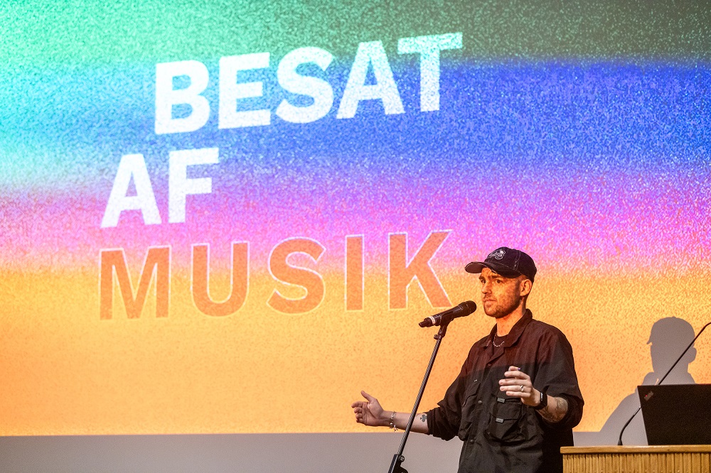 En person står ved en mikrofon og præsenterer sig foran en farverig skærm, der viser ordene "BESAT AF MUSIK".