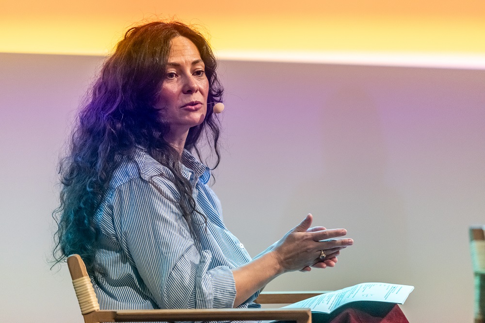 En kvinde med langt mørkt hår sidder på en stol, taler med en headset-mikrofon og holder et stykke papir mod en gradientbaggrund.