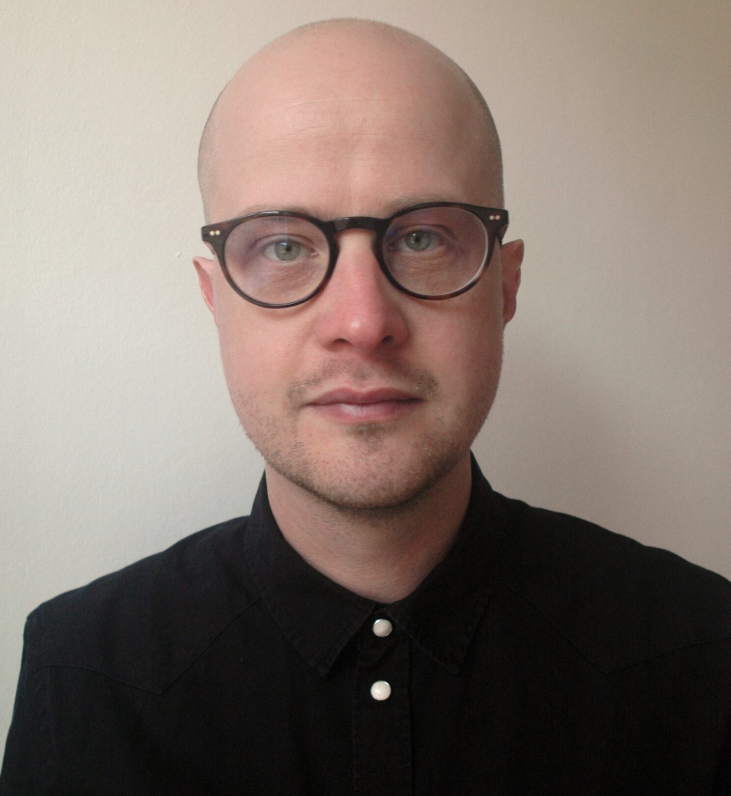 En person med glatbarberet hoved, runde briller og en sort skjorte med krave står foran kameraet på en almindelig lys baggrund.