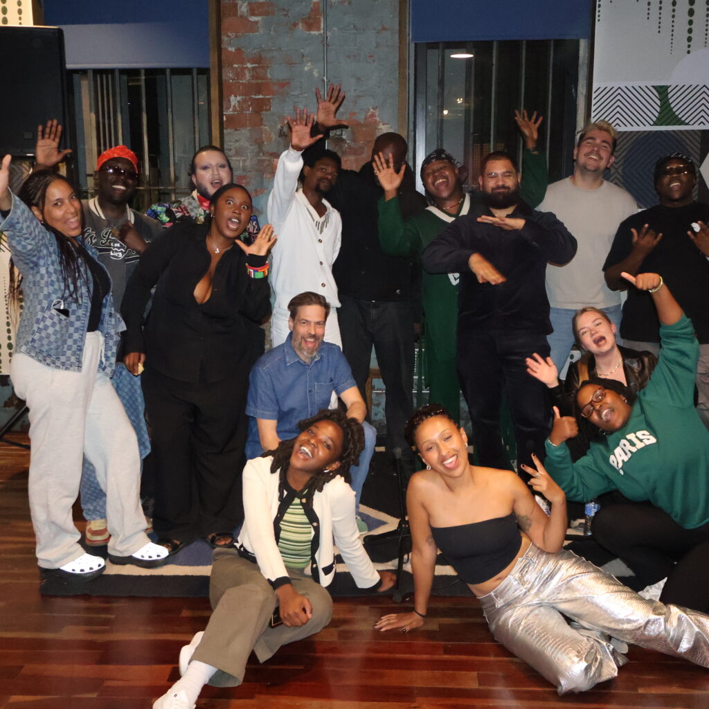 En gruppe mennesker poserer sammen indendørs, smilende og med legende bevægelser, med mønstrede vægge og trægulve i baggrunden.