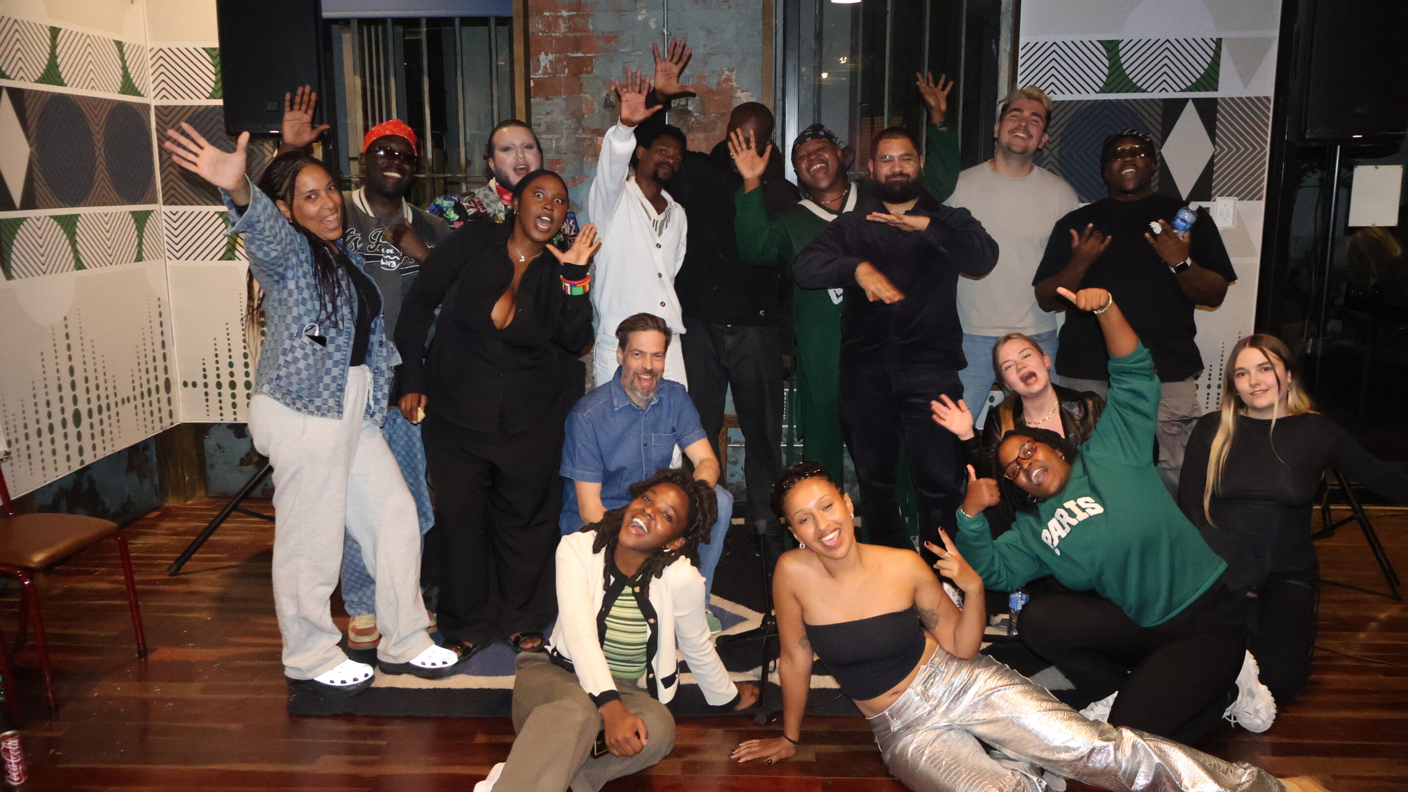 En gruppe mennesker poserer sammen indendørs, smilende og med legende bevægelser, med mønstrede vægge og trægulve i baggrunden.