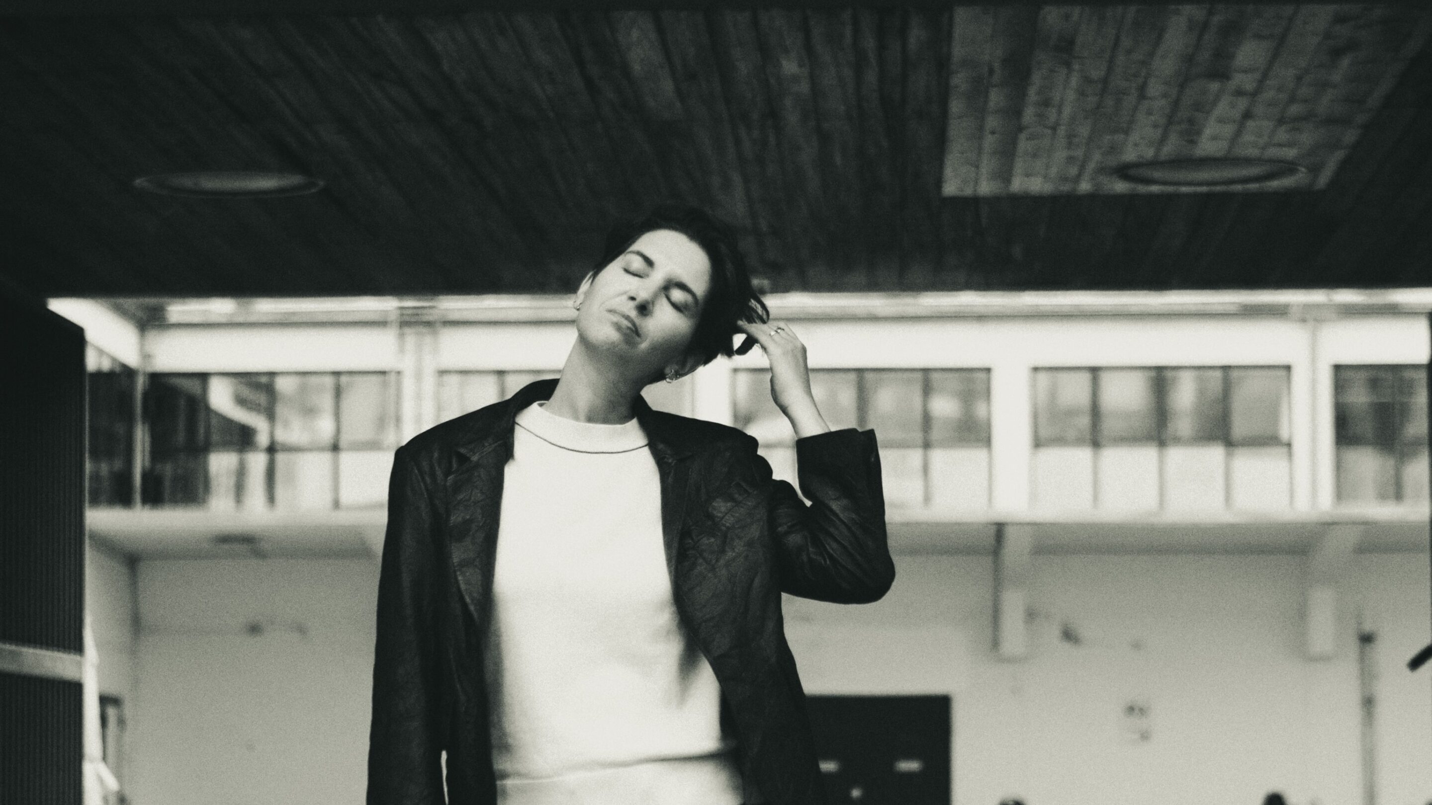 En person står under et træudhæng med lukkede øjne og rører ved sit hoved med en bygning og cykler i baggrunden. Billedet er i sort og hvid.