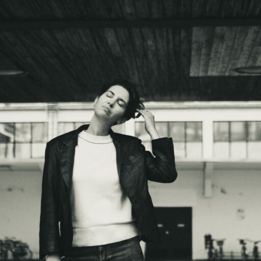 En person står under et træudhæng med lukkede øjne og rører ved sit hoved med en bygning og cykler i baggrunden. Billedet er i sort og hvid.