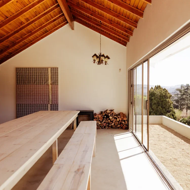 Et langt træbord med bænke står under et hvælvet træloft i et lyst rum med en stak brænde, vægkunst og store glasskydedøre med udsigt over et naturskønt landskab.