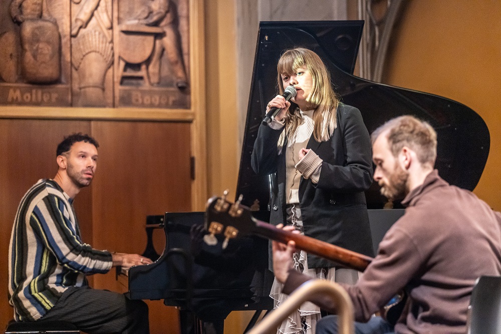 Tre musikere optræder indendørs; en spiller klaver, en synger i en mikrofon, og en spiller guitar. I baggrunden ses et trærelief og et flygel.