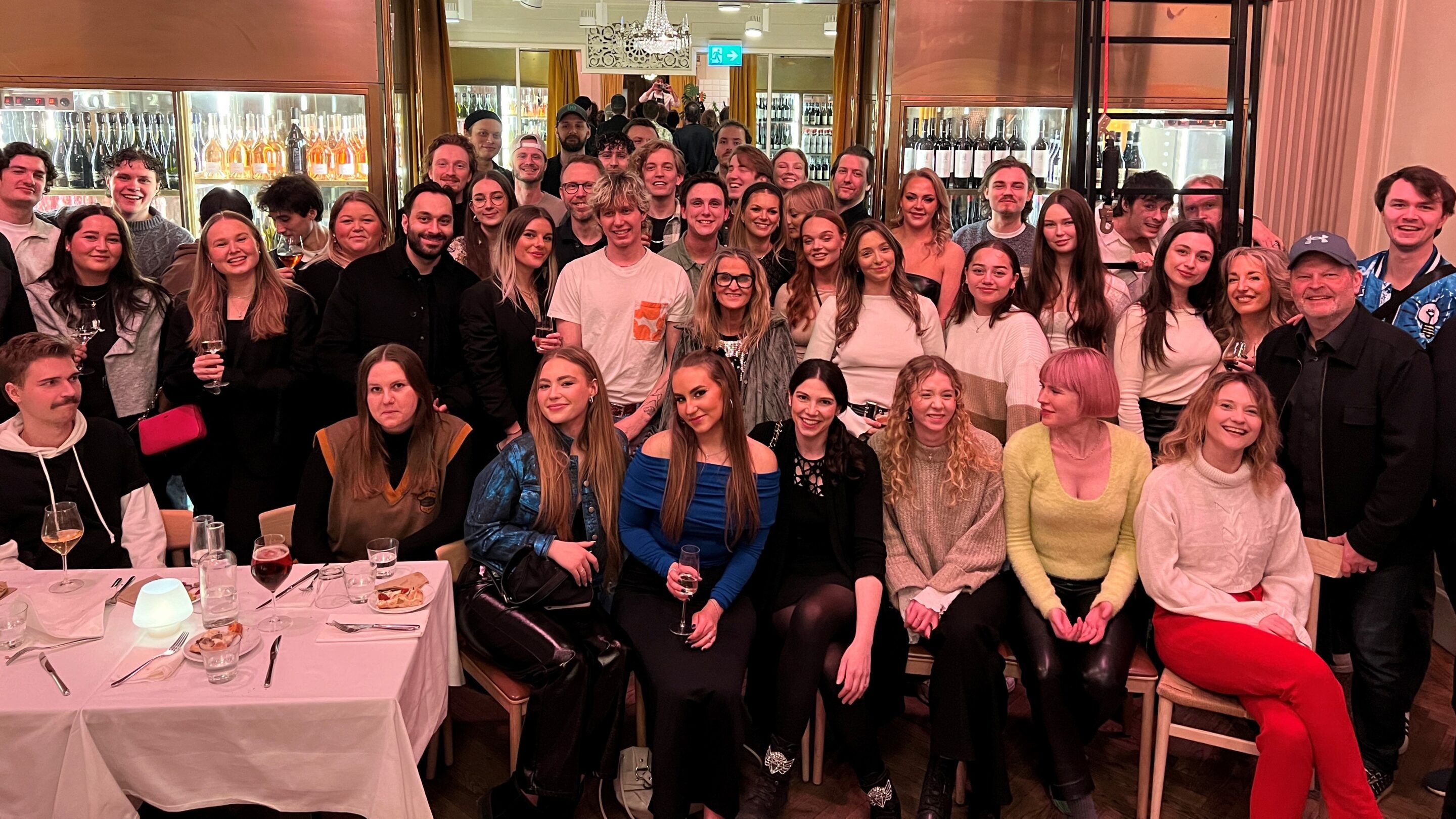 En stor gruppe mennesker poserer og smiler sammen i en restaurant, nogle sidder ved et spisebord, mens andre står bag dem, med glas og flasker synlige i baggrunden.