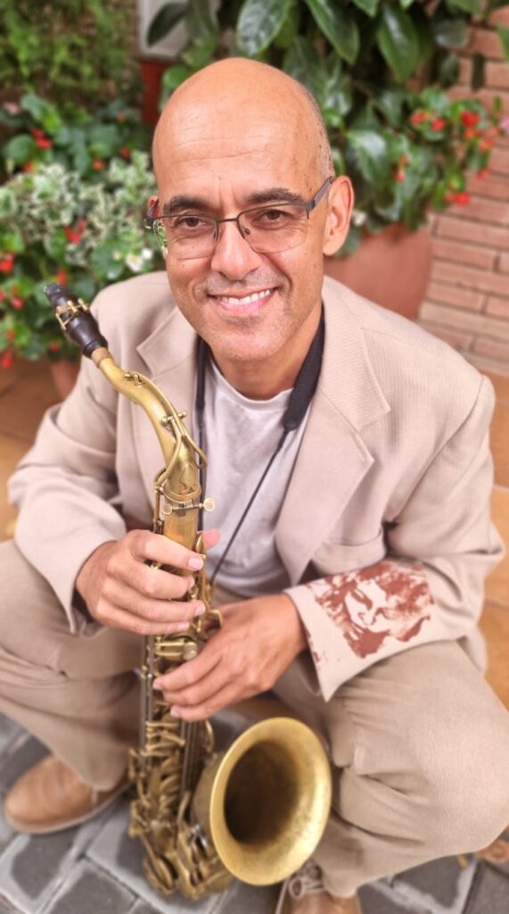 En mand med briller og beige jakkesæt knæler udendørs og smiler til kameraet, mens han holder en saxofon med planter i baggrunden.