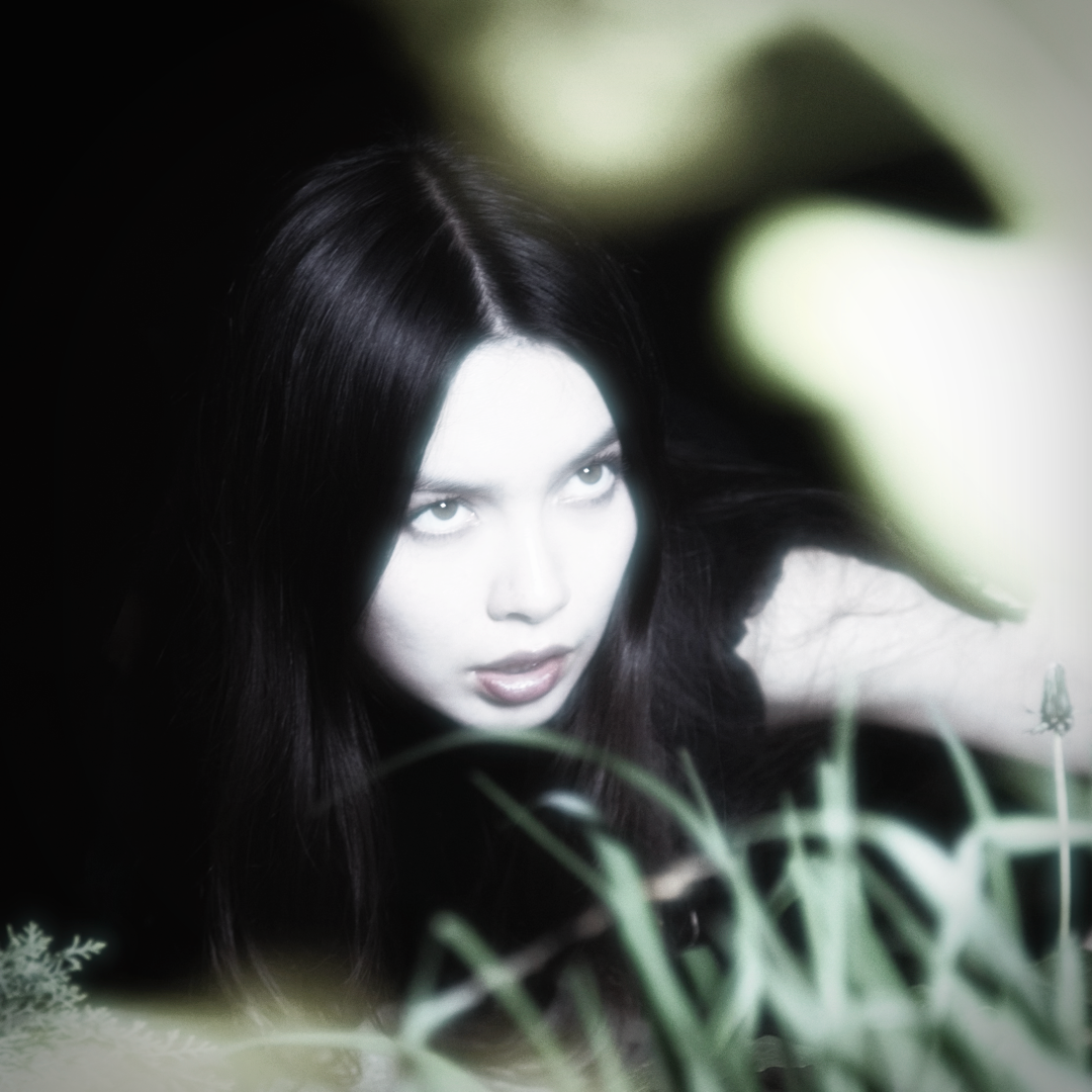 En kvinde med langt mørkt hår kigger op gennem sløret grønt løv, delvist skjult af planter i forgrunden, mod en mørk baggrund.