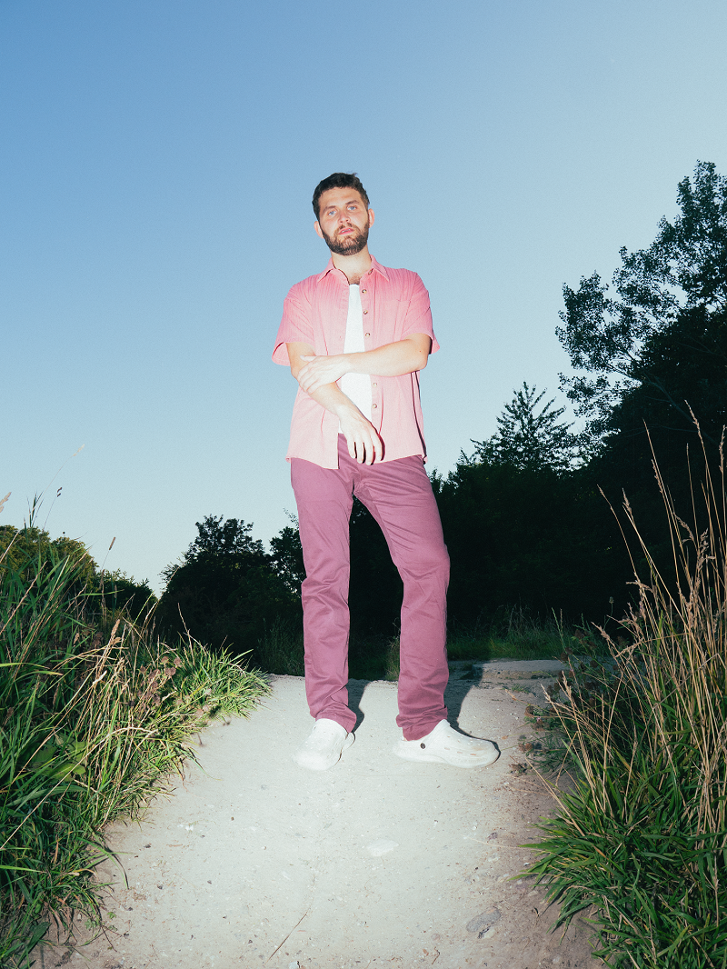 En mand i lyserød skjorte og rødbrune bukser står på en grussti udendørs, omgivet af højt græs og træer, under en klar blå himmel.
