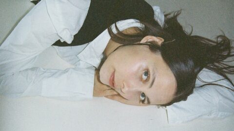 En kvinde med glat mørkt hår og en hvid skjorte i lag med en sort vest ligger på siden, hviler hovedet på armen mod en lys overflade og kigger ind i kameraet.