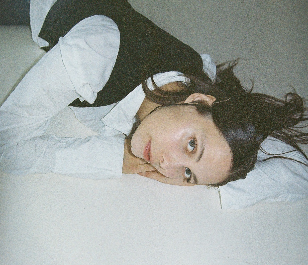 En kvinde med glat mørkt hår og en hvid skjorte i lag med en sort vest ligger på siden, hviler hovedet på armen mod en lys overflade og kigger ind i kameraet.