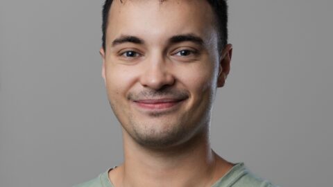 En ung mand med kort mørkt hår og lyst ansigtshår smiler let og er iført en lysegrøn t-shirt på en almindelig grå baggrund.