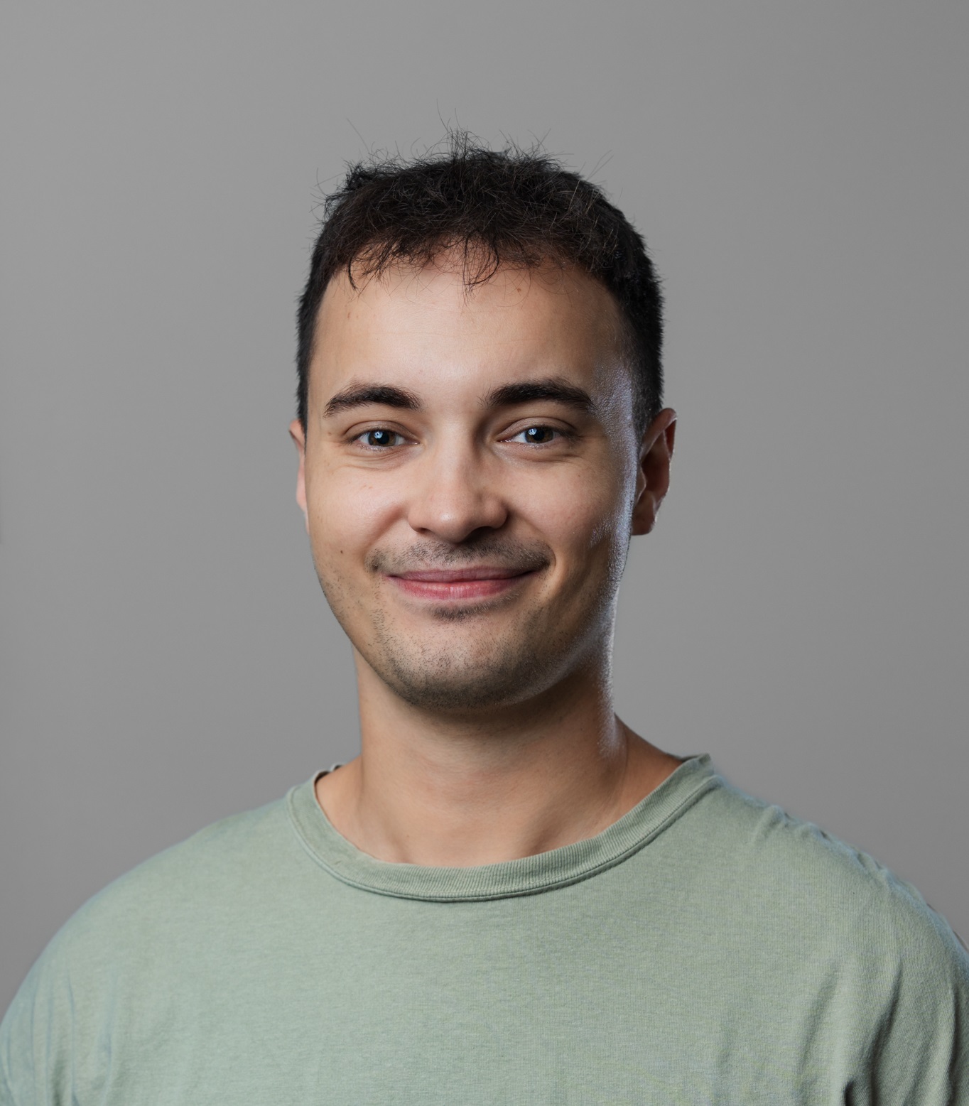 En ung mand med kort mørkt hår og lyst ansigtshår smiler let og er iført en lysegrøn t-shirt på en almindelig grå baggrund.