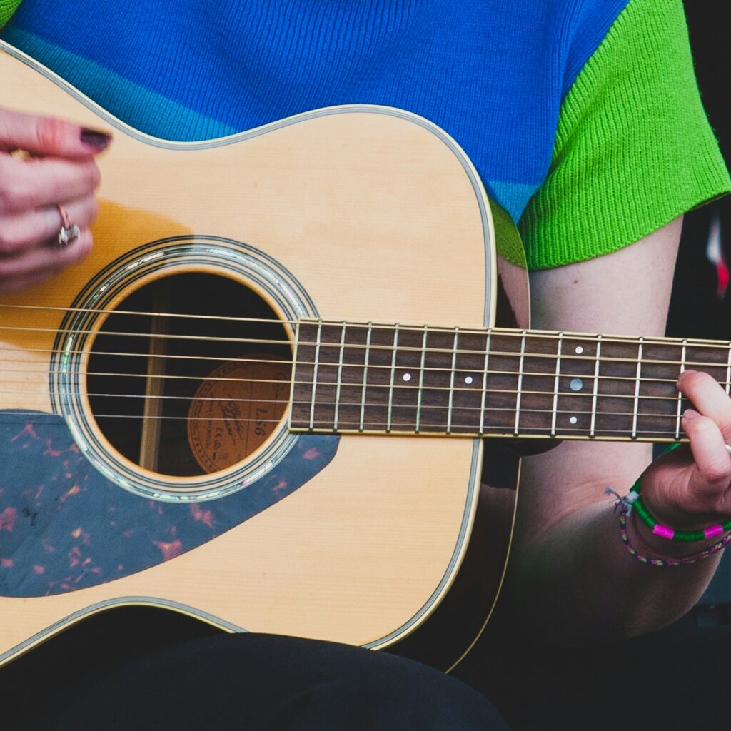 En person iført en grøn og blå skjorte spiller på en akustisk guitar med en capo på halsen og et keyboard i baggrunden.