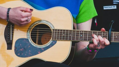 En person iført en grøn og blå skjorte spiller på en akustisk guitar med en capo på halsen og et keyboard i baggrunden.