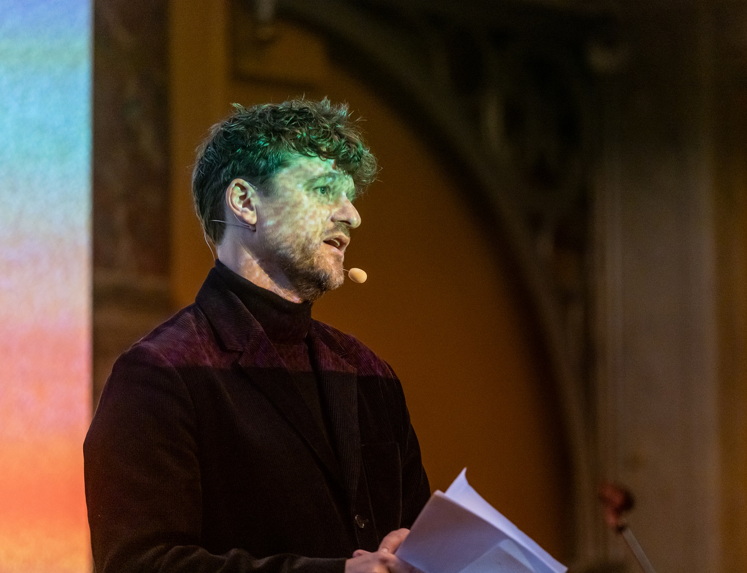 En person med et mikrofonheadset taler, mens han holder papirer, og står indendørs foran en sløret, farverig baggrund.