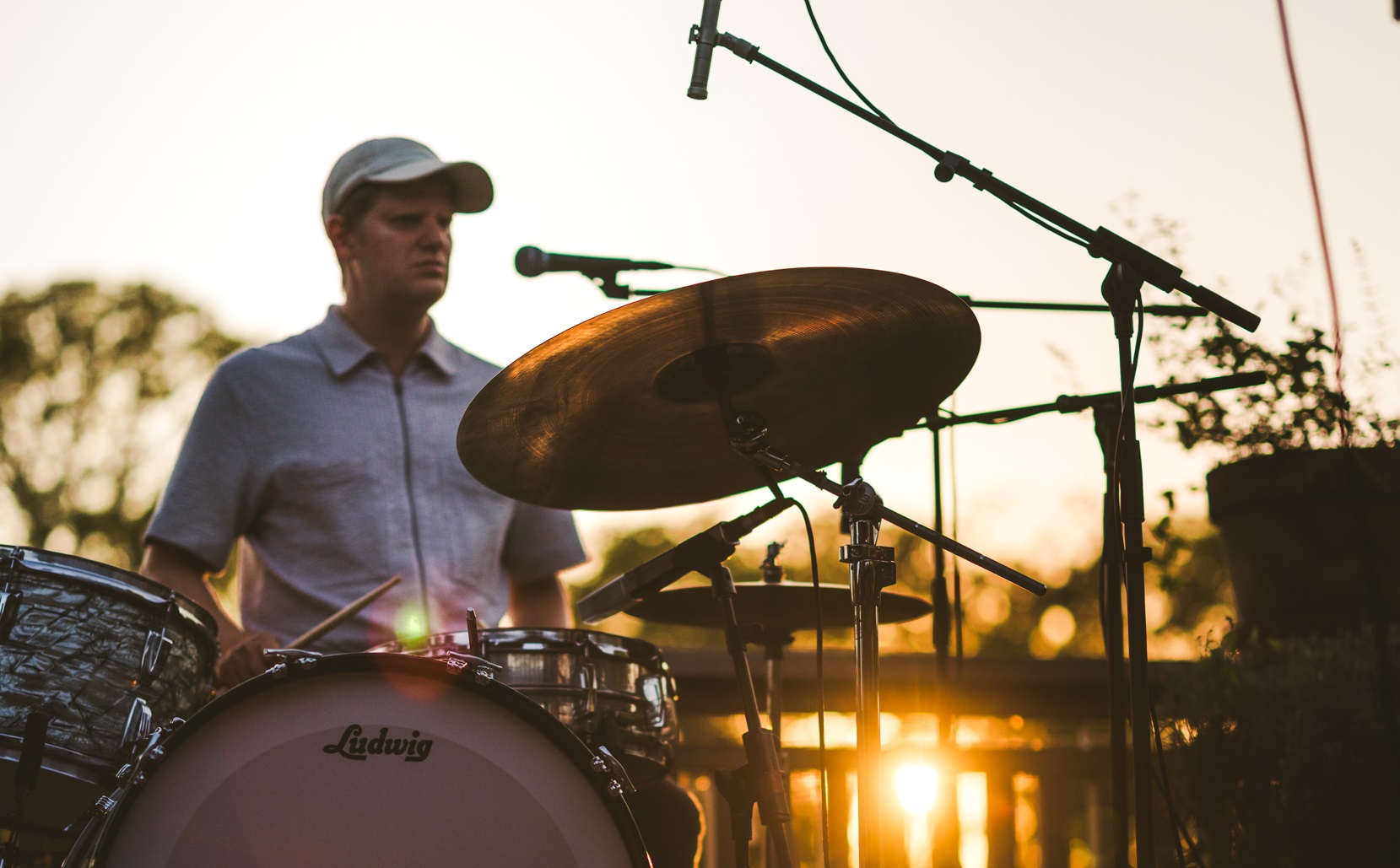 En person spiller på et trommesæt udendørs ved solnedgang med mikrofoner opstillet omkring trommerne og sollys i baggrunden.