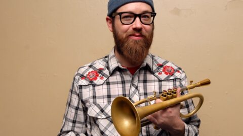 En skægget mand med briller, blå hue og ternet skjorte holder et messinghorninstrument op mod en almindelig beige væg.