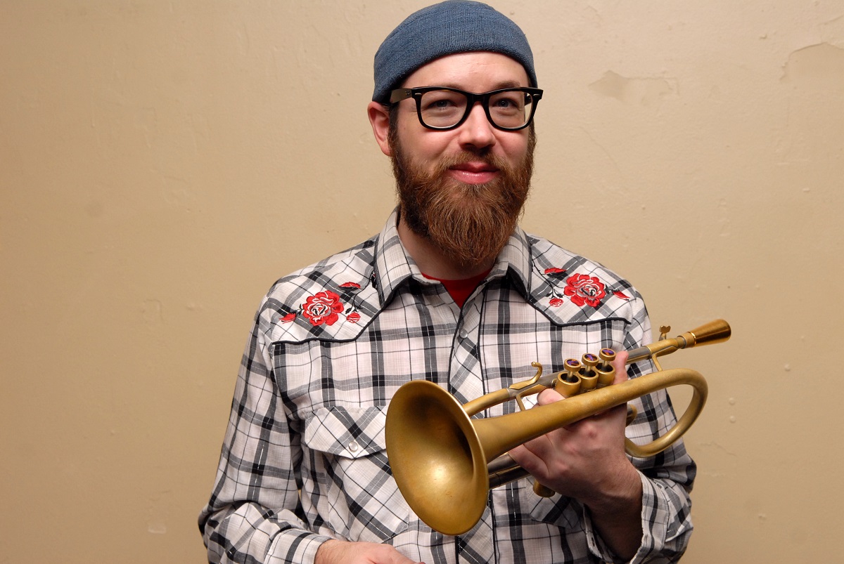 En skægget mand med briller, blå hue og ternet skjorte holder et messinghorninstrument op mod en almindelig beige væg.