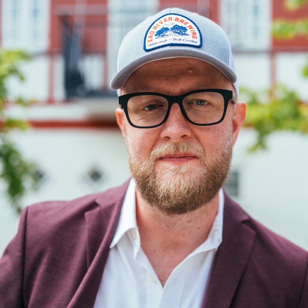 En skægget mand med briller, grå kasket og rødbrun blazer står udendørs foran en bygning med træer i baggrunden.