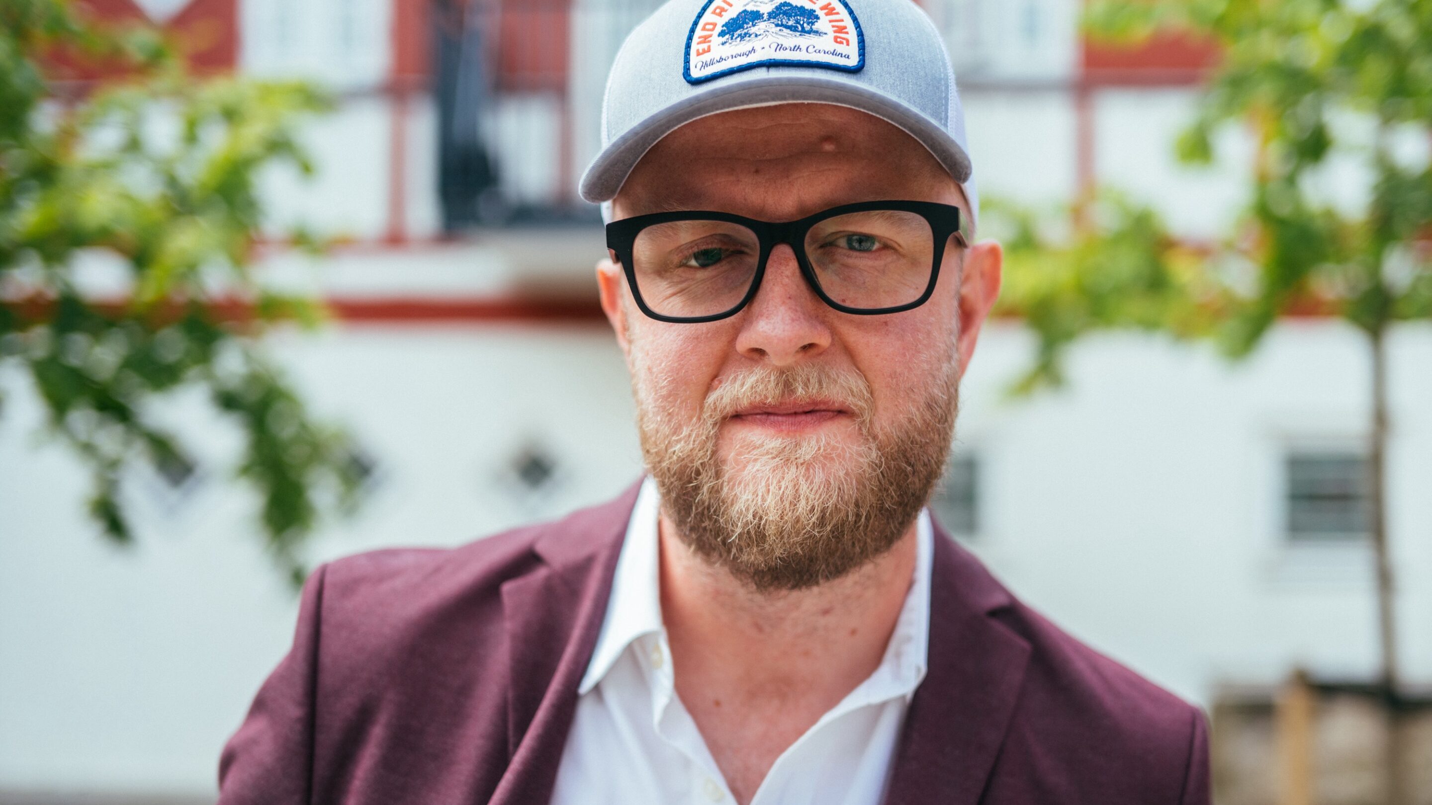 En skægget mand med briller, grå kasket og rødbrun blazer står udendørs foran en bygning med træer i baggrunden.