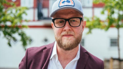 En skægget mand med briller, grå kasket og rødbrun blazer står udendørs foran en bygning med træer i baggrunden.