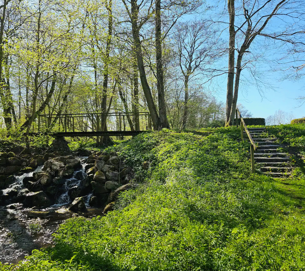En lille træbro går over et stenet vandløb i en solbeskinnet, græsklædt park med stentrapper, der fører op ad en bakke mellem høje træer.