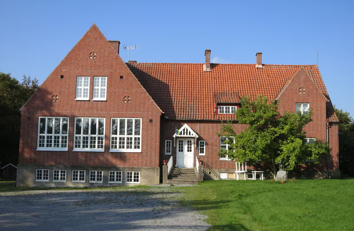 Et toetagers murstenshus med store hvide vinduer, rødt tegltag og en central trappe, der fører til hovedindgangen, ligger på en græsplæne under en klar himmel.