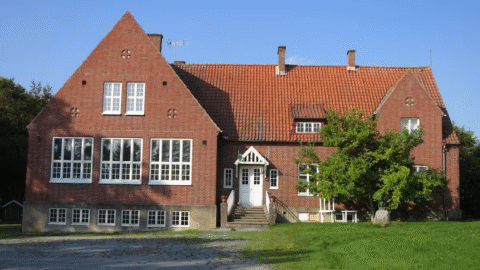 Rødt murstenshus med store vinduer, hvide kanter og et stejlt tegltag, omgivet af græs og træer under en klar blå himmel.