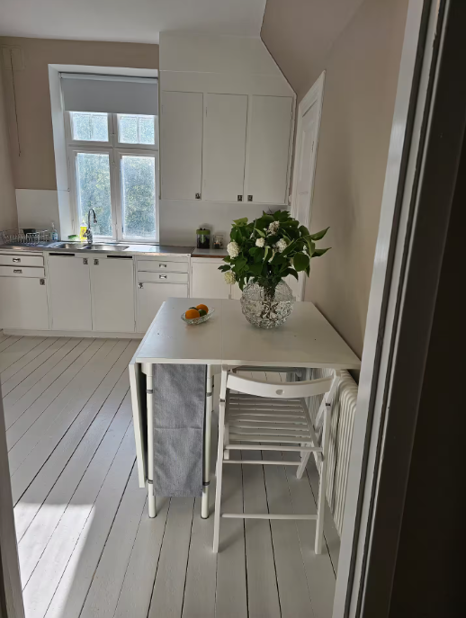 Et lille, hvidt køkken med et sammenklappeligt bord, en vase med blomster, en stol og appelsiner på bordet ved siden af et vindue med sollys, der strømmer ind.