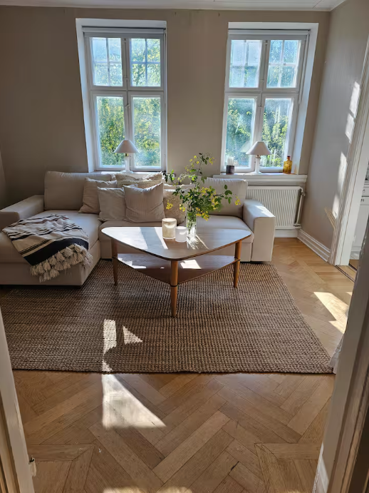 En solbeskinnet stue med en beige sofa, et sofabord i træ med blomster, et vævet tæppe og to vinduer, der lukker naturligt lys ind.