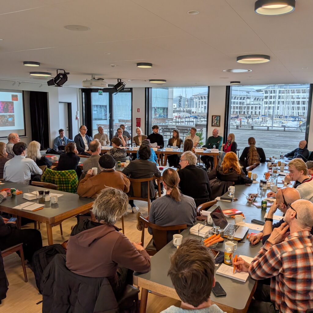 En gruppe mennesker sidder omkring borde i et konferencelokale og lytter til talere ved et langt bord; en præsentation vises på en skærm foran.