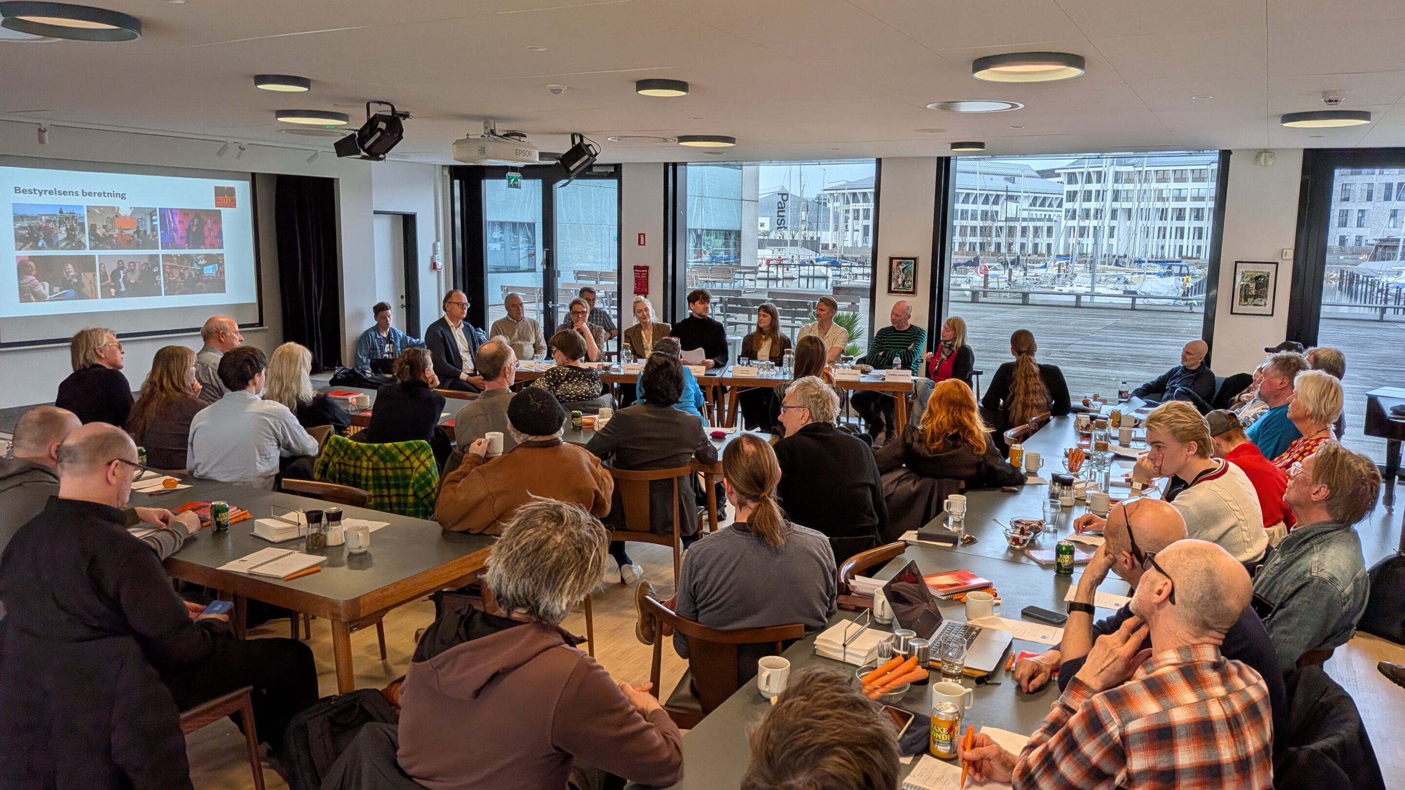 En gruppe mennesker sidder omkring borde i et konferencelokale og lytter til talere ved et langt bord; en præsentation vises på en skærm foran.