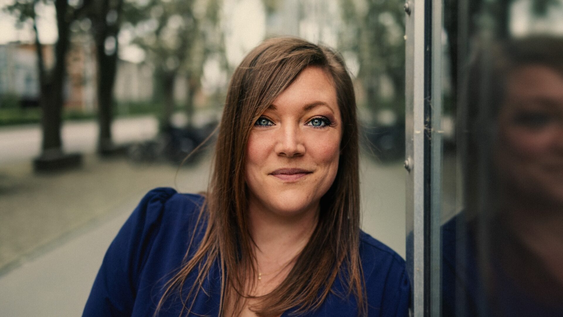 En kvinde med glat, brunt hår og en blå top står udendørs og læner sig op ad en reflekterende glasoverflade med træer og bygninger i baggrunden.