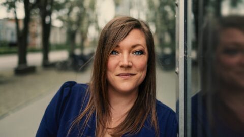 En kvinde med glat, brunt hår og en blå top står udendørs og læner sig op ad en reflekterende glasoverflade med træer og bygninger i baggrunden.
