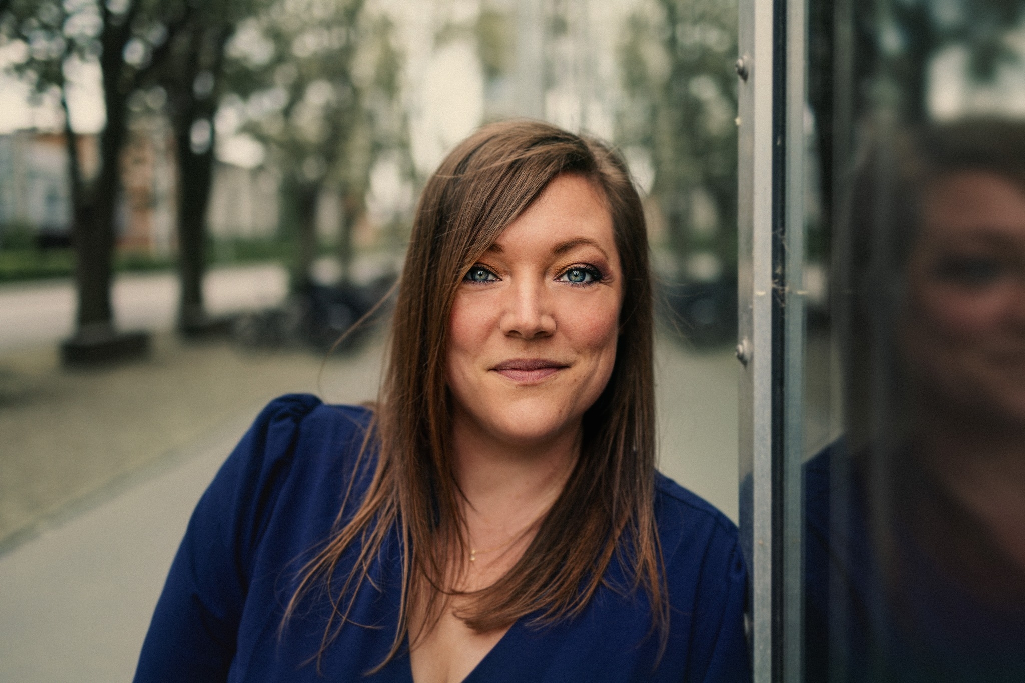 En kvinde med glat, brunt hår og en blå top står udendørs og læner sig op ad en reflekterende glasoverflade med træer og bygninger i baggrunden.