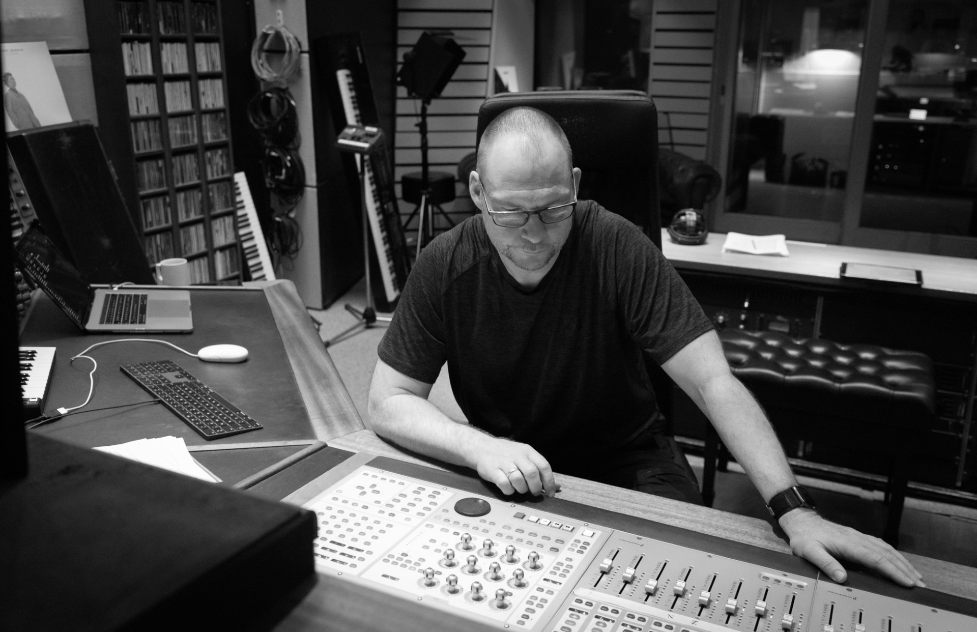 En person sidder ved en stor lydmixer i et indspilningsstudie og fokuserer på kontrollerne. I baggrunden ses tastaturer og hylder med medier.