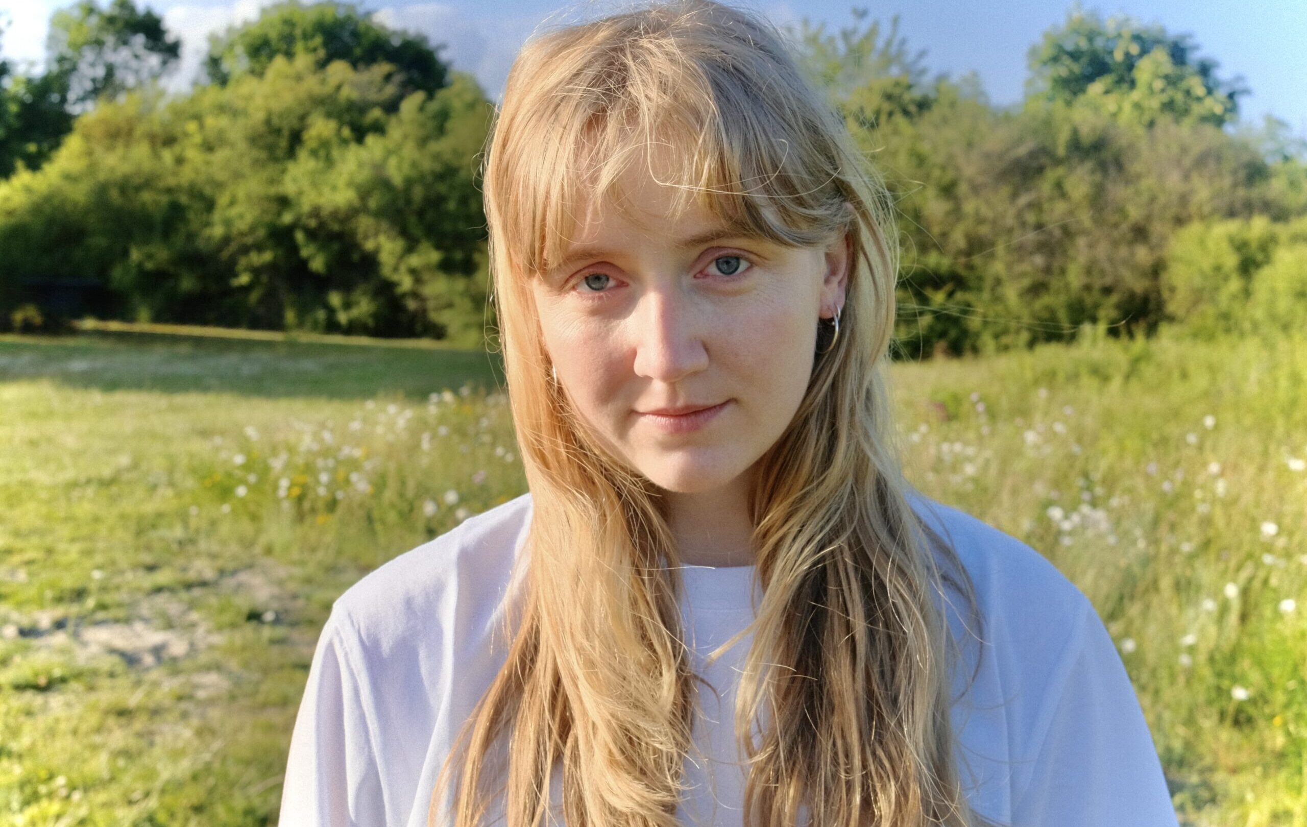 En ung kvinde med langt blondt hår står udendørs på en græsmark og kigger ind i kameraet med træer og blå himmel i baggrunden.