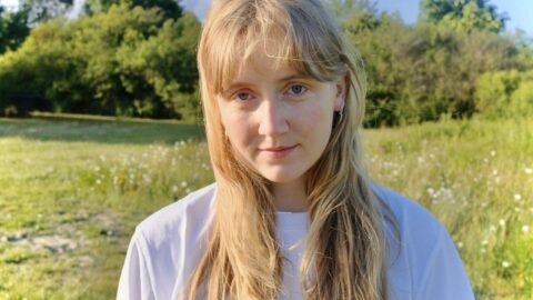 En ung kvinde med langt blondt hår står udendørs på en græsmark og kigger ind i kameraet med træer og blå himmel i baggrunden.