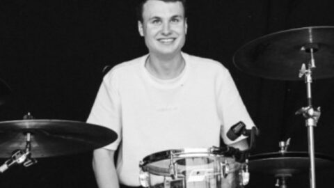 En person i en lys t-shirt sidder smilende ved et trommesæt med en mørk baggrund bag sig.