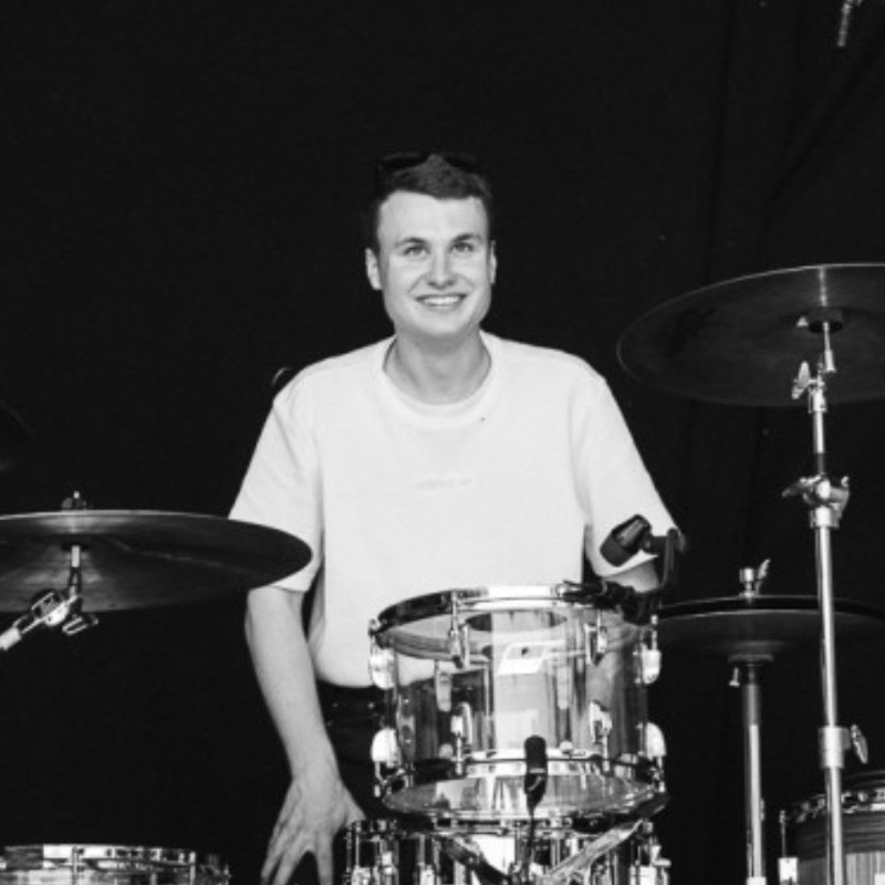 En person i en lys t-shirt sidder smilende ved et trommesæt med en mørk baggrund bag sig.