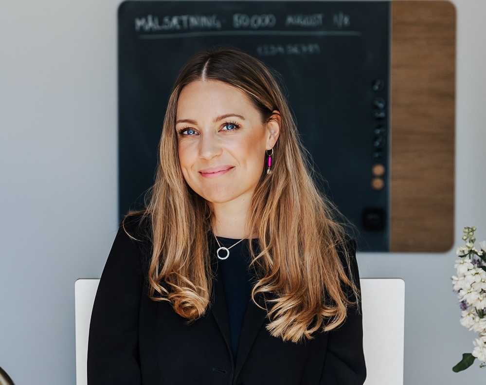 En kvinde med langt blondt hår og sort tøj sidder foran en kridttavle med skrift og tal.