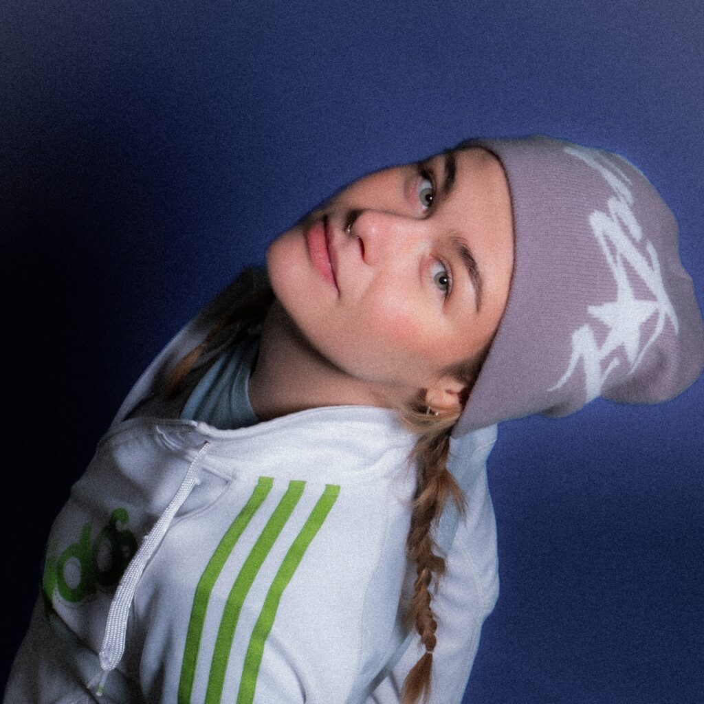 Kvinde i hvid hættetrøje med grønne striber og lyslilla hue, der lægger hovedet på skrå og kigger ind i kameraet mod en ensfarvet lilla baggrund.